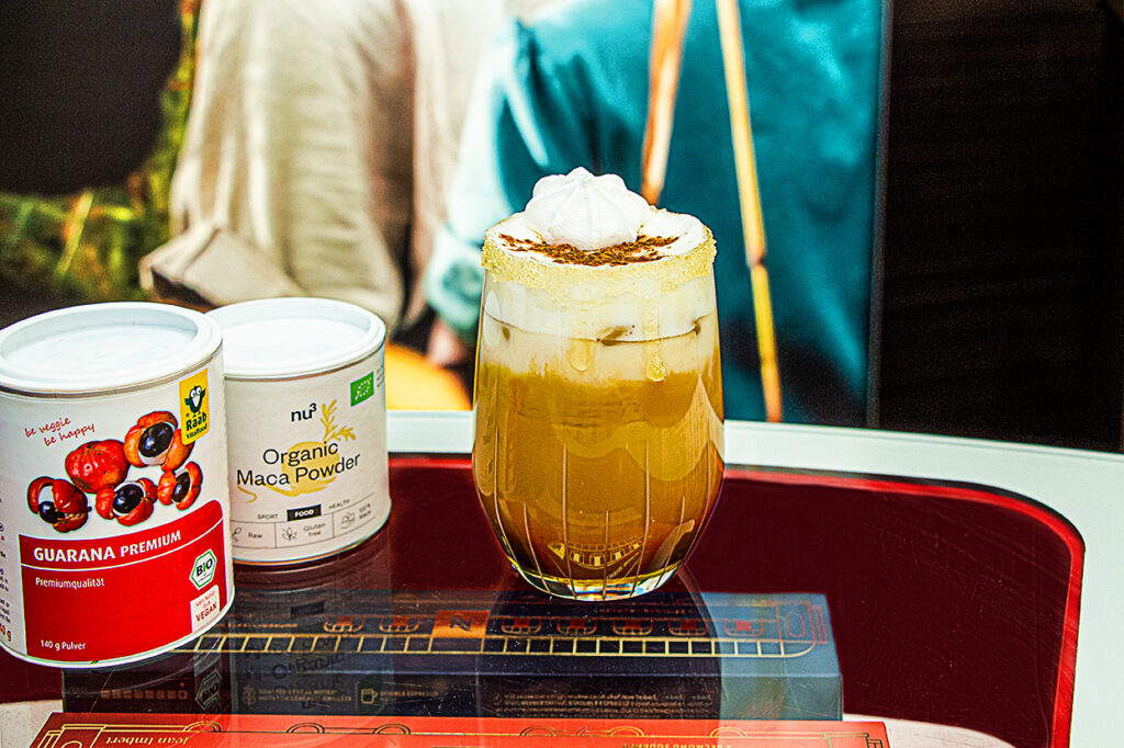 „Nahaufnahme eines funktionalen Kaffeecocktails im Glas mit Milchschaum, Zimt und Zuckerkruste neben Dosen mit Guarana-Premium- und Bio-Maca-Pulver auf rotem Tablett.“