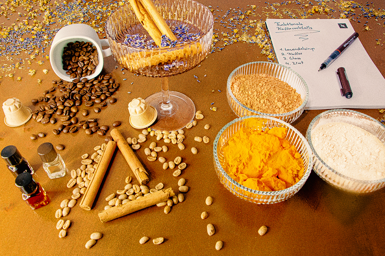 „Flatlay mit Zutaten für funktionale Kaffeecocktails: Lavendelsirup im Coupéglas, verstreute Kaffeebohnen, Kaffeekapseln, Zimtstangen, bunte Gewürz- und Pflanzenpulver in Glasschalen sowie ein Notizblock mit Rezeptliste auf goldenem Untergrund.“