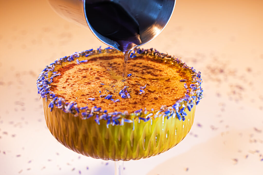 „Nahaufnahme eines funktionalen Kaffeecocktails im Coupéglas, dessen Rand mit getrockneten Lavendelblüten garniert ist, während duftender Lavendelsirup aus einem Milchkännchen in das Glas fließt und zuvor Zimt darüber gestreut wurde.“