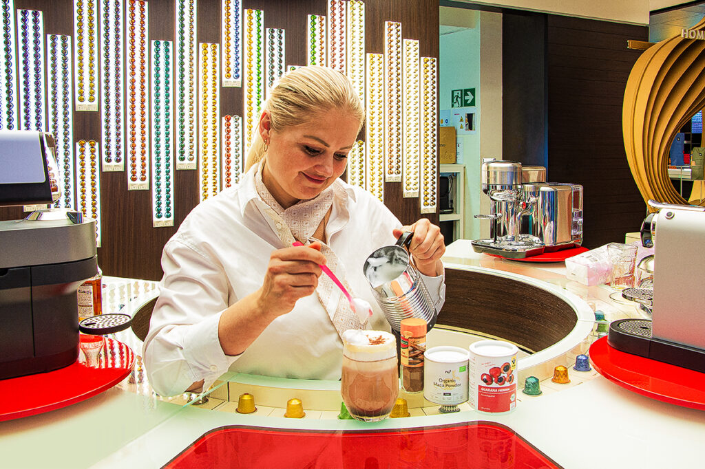 „Barista an einer Nespresso-Station rührt Milchschaum mit einem Löffel in einen Glas-Kaffeecocktail, umgeben von Nespresso-Kapseln, Maca- und Guarana-Pulvern sowie Kaffeemaschinen.“