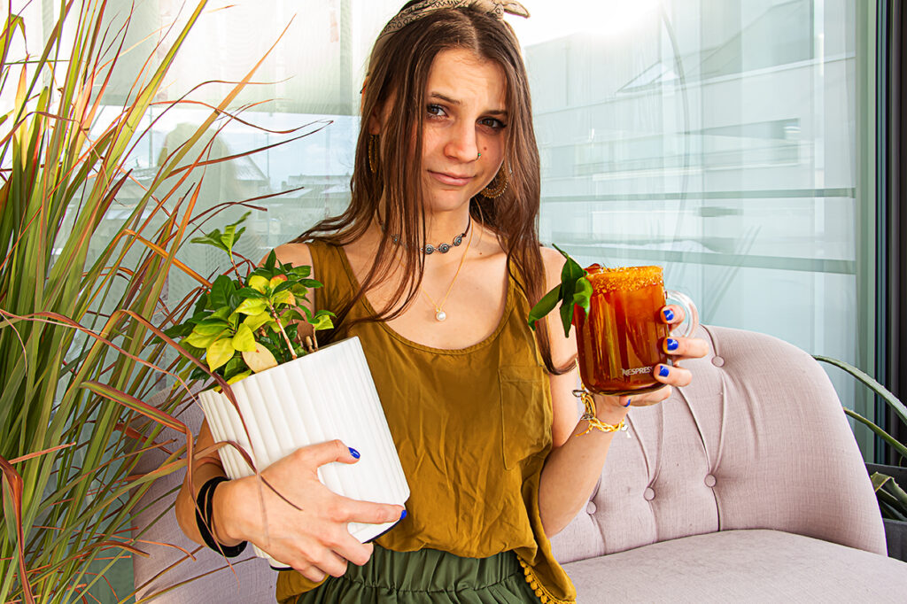 „Junge Frau im Wohnzimmerhimmel mit blauer Nägeln hält in der einen Hand einen Topf mit grüner Zimmerpflanze und in der anderen einen Kaffeebecher mit Nespresso-Logo, gefüllt mit einem dunkelorangenen Kaffeecocktail, umgeben von pflanzlichen Dekoelementen.“