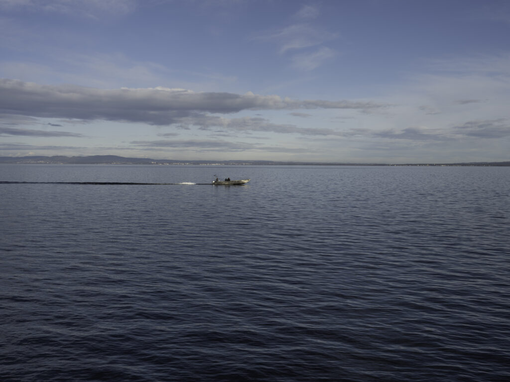 Kleines Motorboot mit zwei Personen fährt über eine weite, ruhige Wasserfläche unter leicht bewölktem Himmel; im Hintergrund ist eine entfernte Uferlinie zu sehen.