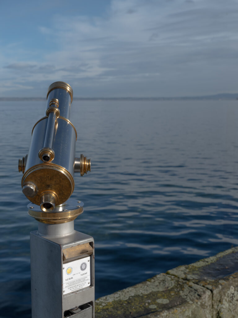 Nahaufnahme eines münzbetriebenen Besucher-Fernrohrs mit goldenen und silbernen Metallteilen, montiert auf einem grauen Sockel mit Münzeinwurf („1 × 2 EUR“), vor dem ruhigen Wasser eines Sees und einem leicht bewölkten Himmel.
