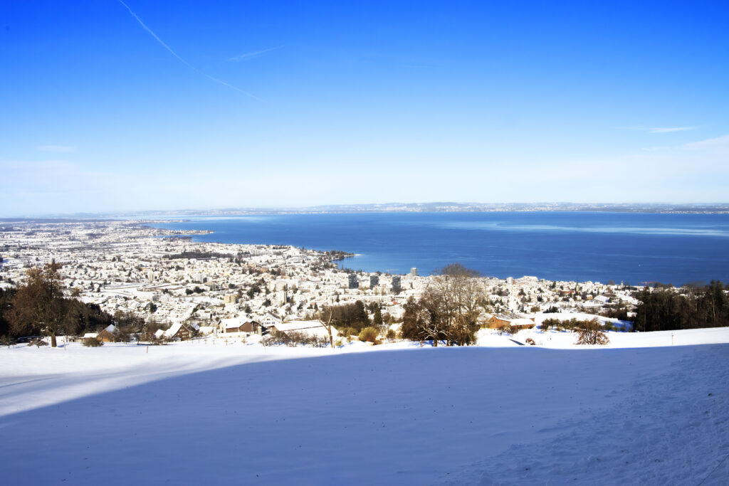 Verschneiter Hang im Vordergrund, darunter eine Stadt mit Häusern und Bäumen am Ufer eines großen, tiefblauen Sees; im Hintergrund erstreckt sich das offene Wasser und ein klarer, strahlend blauer Himmel.