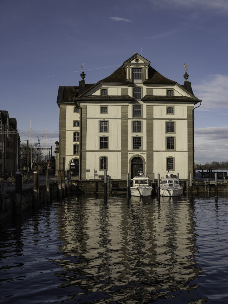Dreistöckiges klassizistisches Gebäude mit hohem, ziegelgedecktem Mansarddach und der Aufschrift „Museum Galerie“ über dem Tor; es steht direkt am Ufer, vor ihm liegen zwei weiße Motorboote, und das Gebäude spiegelt sich im ruhigen Wasser.