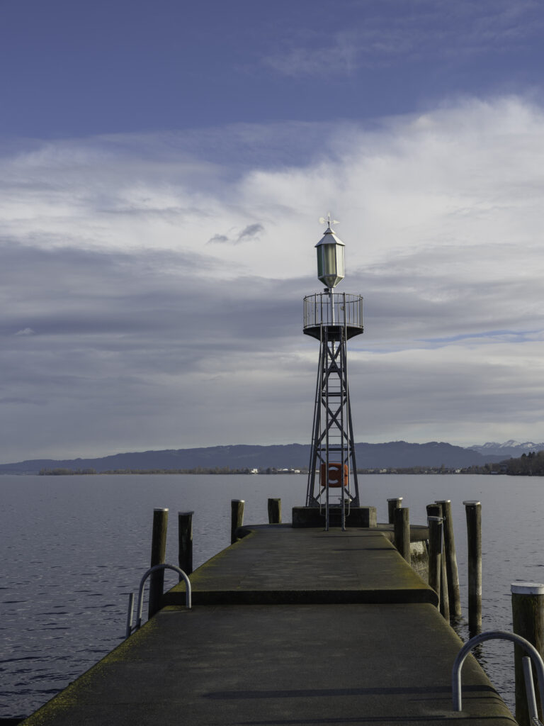 Ein steinerner Bootssteg, der auf einen kleinen eisernen Peilturm mit rundem Geländer und einer grünen Navigationslaterne führt; links und rechts stehen Holzpfähle im ruhigen Wasser eines großen Sees, im Hintergrund Berge und bewölkter Himmel.